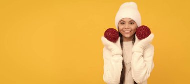 Kışlık şapkalı ve eldivenli neşeli çocuk sarı arka planda dekoratif xmas topları tutuyor, xmas. Noel çocuğu, stüdyo çocuğu kışın fotokopi çekilen portresi