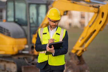 İnşaat alanında mimar. Modern ev inşaatında kask ve takım elbiseli mimar. Güvenlik yeleği ve takım elbisesi olan bir mimar. Evin arka planında duran kendine güvenen mimar.