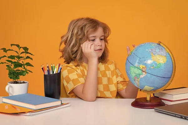 School and education concept. Portrait of cute child school boy looking at globe during geography lesson isolated on yellow studio background. Back to school