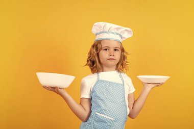 Funny kid chef cook with kitchen plate, studio portrait. Kid chef cook prepares food on isolated studio background. Kids cooking. Teen boy with apron and chef hat preparing a healthy meal