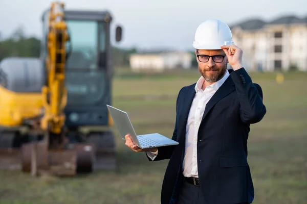 Architect at a construction site. Architect man in helmet and suit at modern home building construction. Architect with a safety vest and suit. Confident architect standing at house background