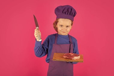 Child cook hold cutting board with meat beef steak and knife. Teen boy with apron and chef hat preparing a healthy meal