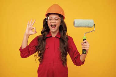 Excited teenager girl. Painter in workshop. Renovation and repair. Teen girl in helmet with painting brush. Child in hard hat. Kid builder painter on construction site