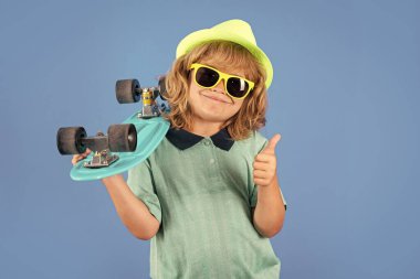 Studio portrait of stylish cute young child boy with skateboard isolated against blue background. Kid skater. Fashion kid. Stylish little child wearing a summer clothes, sunglasses and hat