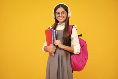 Schoolgirl, teenage student girl in headphones hold books on yellow isolated studio background. School and music education concept. Back to school. Happy face, positive and smiling emotions.