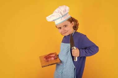 Kid cook hold cutting board with meat beef steak and knife. Cooking, culinary and kids. Little boy in chefs hat and apron on studio isolated background