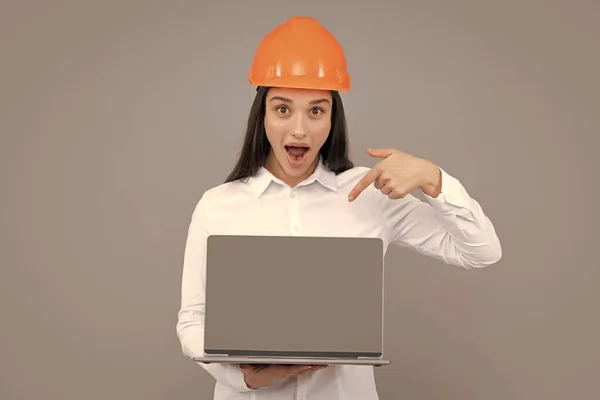 Surprised shocked excited business woman with laptop computer, portrait on gray. Young woman in helmet hold laptop computer on gray background. Architect businesswoman