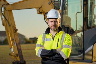 İnşaat alanında inşaat için kazı yapan bir inşaat işçisi. Binaların arka planında inşaat işçisi olan bir makine traktörü. Kazı işçisi. İnşaat alanında miğferi olan bir inşaatçı