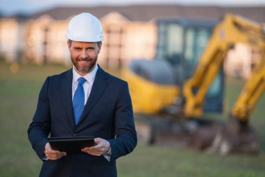 İnşaat alanında kask ve takım elbiseli bir inşaat yöneticisi. İnşaat müdürü ya da yönetici evin önünde şapka takıyor. Kazıcının yanındaki inşaat müdürü. Yenileme