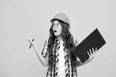 Shocked surprised teenager girl. Teenager child painter in helmet with clipboard. Child in hard hat. Kid engineer on construction site. Renovation and repair