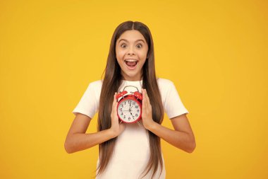 Saati, zamanı ve son teslim tarihi olan genç bir kızın portresi. Stüdyo çekimi sarı arkaplanda izole edildi. Heyecanlı bir genç, şaşkın ve neşeli duygular içinde