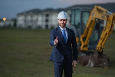 İnşaat alanında kask ve takım elbiseli bir inşaat yöneticisi. İnşaat müdürü ya da yönetici evin önünde şapka takıyor. Kazıcının yanındaki inşaat müdürü. Yenileme