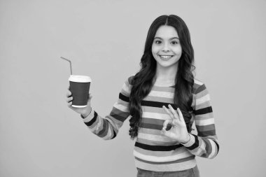 Child with coffee or tea cup isolated on yellow studio background. Teenage girl with take away beverage. Happy teenager, positive and smiling emotions of teen girl