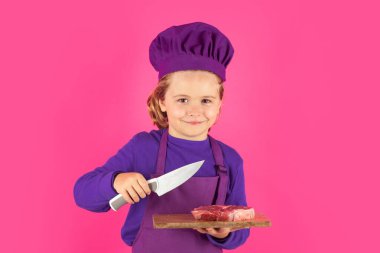 Çocuk aşçı et bifteği ve bıçakla kesme tahtasını tutuyor. Üniformalı çocuk. Aşçı çocuk pembe arka planda izole edilmiş. Aşçı olmak için sevimli bir çocuk. Aşçı şapkası giymiş bir çocuk.