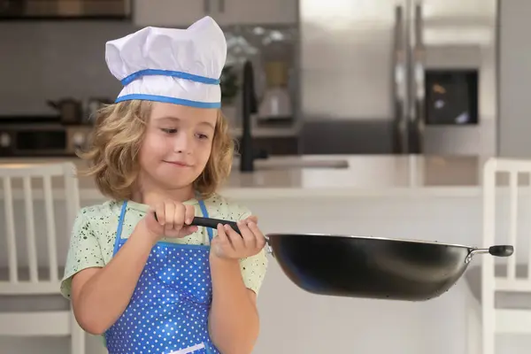 Kid chef cook cookery with pan in kitchen. Children cooking in the kitchen. Funny kid chef cook cookery at kitchen. Chef kid boy making healthy food. Portrait of little child in chef hat