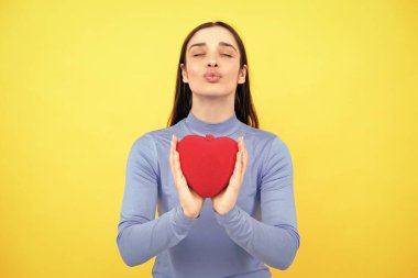 Mutlu kadın kırmızı kalbi tutar. İzole edilmiş sarı arka planda poz veren güzel bir kadın model. Neşeli, pozitif, tatlı, yumuşak kalpli bir kız.