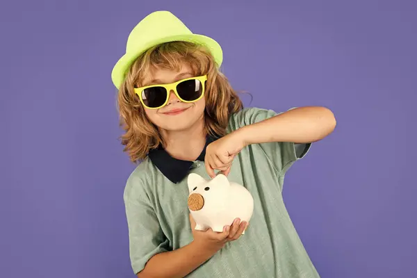 Saving money and piggy bank concept. Portrait of a little boy putting money on a moneybox. Child saving money in a piggybank on blue background