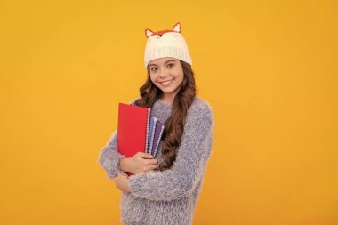 Çalışma kitabını tutan çocuğun portresi. Olumlu duygularını ifade et. Kış modası. Şapkalı, kıvırcık saçlı, gülümseyen çocuk. Okula dönüyorum. Sarı arka planda örgü elbiseli genç kız..