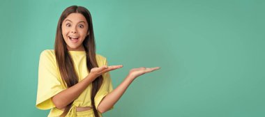 surprised kid presenting product on blue background with copy space, here. Child face, horizontal poster, teenager girl isolated portrait, banner with copy space
