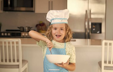Çocuk aşçı yemek tabağıyla yemek yapıyor. Eğlenceli çocuklar mutfağı. Üniformalı aşçı şapkası ve önlük takan komik aşçı çocuk mutfakta yemek pişirdi. Çocuklar yemek hazırlıyor, mutfakta kurabiye pişiriyor.