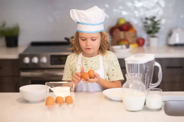 Child chef cook with eggs. Funny little kid chef cook wearing uniform cook cap and apron cooked food in the kitchen. Kids are preparing the dough, bake cookies in the kitchen