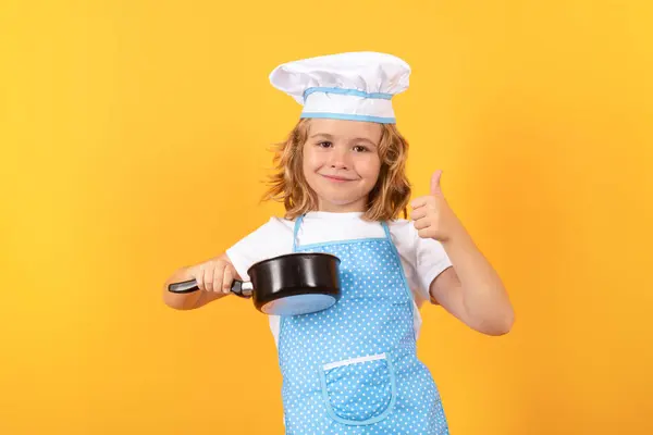 Kid chef cook with cooking pot stockpot. Kid chef cook prepares food on isolated studio background. Kids cooking. Teen boy with apron and chef hat preparing a healthy meal