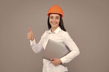 Young woman in helmet hold laptop computer on gray background. Architect businesswoman in helmet thinking, young caucasian female in work as constructor engineer