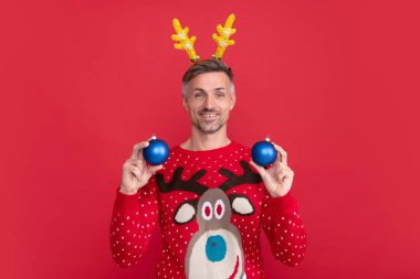 Kış süveteri ve parti boynuzlarıyla gülen adam. Adam kırmızı arka planda Xmas dekorasyon topları tutuyor. Mutlu yıllar. Mutlu Noeller..