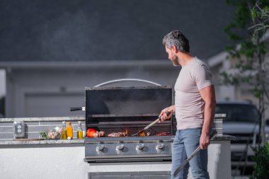 Barbekü şefi. Barbekü için önlüklü orta yaşlı İspanyol bir adam. Kızartma ve ızgara yemek. Adam mutfak aletlerini barbeküde tutuyor. Açık havada et kızartıyoruz. Barbekü ve ızgara. Arka bahçede et pişiriyorum.