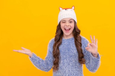 Sarı arka planda örgü giyen genç bir kız. Kalın giysiler giyen çocuk portresi. Olumlu duygularını ifade et. Kış modası. Göz kırpan çocuk fotokopi alanında ürün sunuyor. Kadın model..