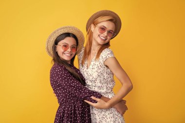 Anne ve kızı sarı arka planda hasır şapkayla gülümsüyor..