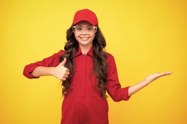 Tulum giyip, şapka takıp gözlük takan genç bir işçi. Stüdyo fotoğrafı sarı arka planda izole edilmiş. Gösterme ve gösterme kavramı