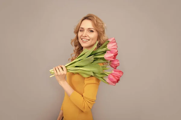 winking cheerful lady hold flowers for spring holiday on grey background.