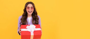 Güneş gözlüklü ve takım elbiseli son moda çocuk sarı arka planda hediye kutusunu tutuyor. Doğum günü hediyesi olan genç bir kız, yatay poster. Kopyalama alanı olan başlık