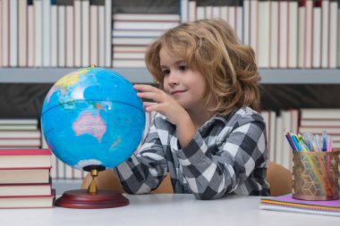 School boy looking at globe in library, geography lesson. School child student learning in class, study english language at school. Elementary school child. Portrait of funny pupil learning