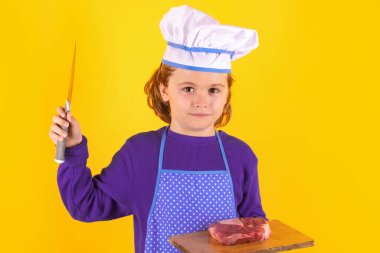 Çocuk aşçı et bifteği ve bıçakla kesme tahtasını tutuyor. Aşçı çocuk, stüdyo resmi. Çocuklar yemek yapıyor. Önlüklü ve aşçı şapkalı çocuk.