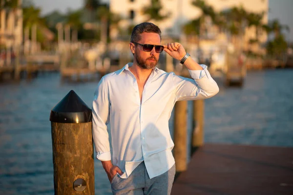 Rich fashion business man dreaming and thinking of yacht. Well-dressed fashion man in fashionable eyeglasses and shirt posing near yacht. Male model in yacht club. Portrait of stylish man. Male