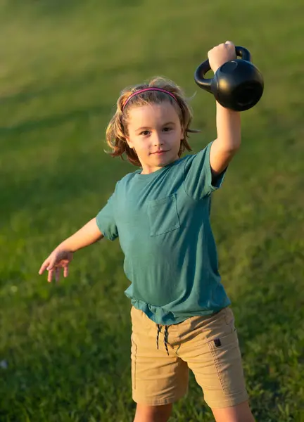 孩子们在外面的后院提起水壶铃铛 可爱的小男孩用水壶鼓起胳膊肌肉 有哑铃的健康儿童 免版税图库图片