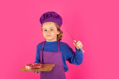 Çocuk aşçı et bifteği ve bıçakla kesme tahtasını tutuyor. Çocuk fırın üniforması giyiyor ve aşçı şapkası takıyor. Mutfakta yemek hazırlıyor, stüdyo fotoğrafı çekiyor. Aşçılık, aşçılık ve çocuk maması konsepti.