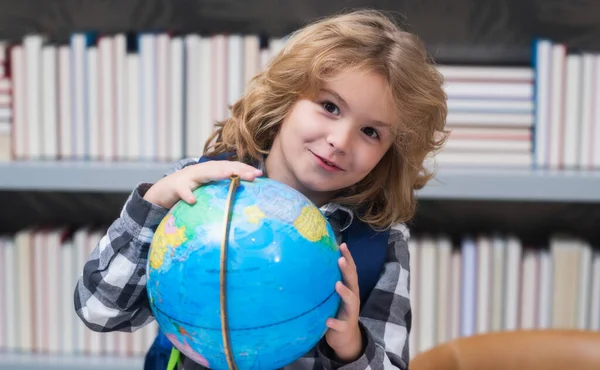 School kid pupil looking at globe in library at the elementary school. World globe. School kid 7-8 years old with book go back to school. Little student. Education concept