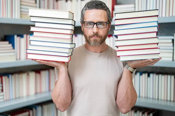 Portrait of funny teacher with book in library classroom. Handsome teacher in university library. Funny teacher hold many books. Crazy teacher with books. Excited teachers in school book library