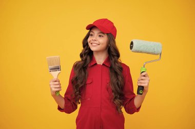 Happy smiling builder in building uniform and cap. Painter with painting brush tool or paint roller. Worker isolated on yellow background. Kids renovation concept