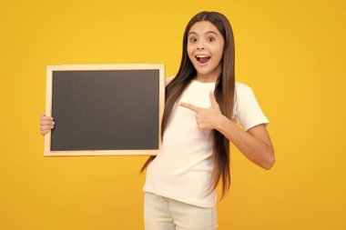 Okulun boş tahtasını tutan liseli genç kız sarı arka planda izole edilmiş. Genç bir kız öğrencinin portresi. Heyecanlı bir genç, şaşkın ve neşeli duygular içinde