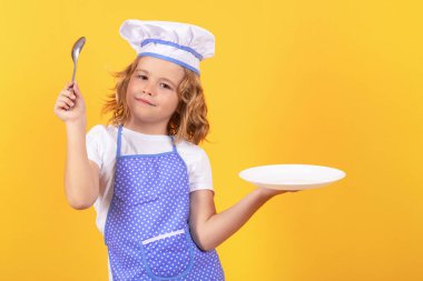 Mutfak tabağı ve kaşıkla komik aşçı çocuk, stüdyo portresi. Aşçı üniformalı küçük bir çocuğun portresi. Şef çocuk stüdyo arka planında izole edilmiş. Aşçı olmak için sevimli bir çocuk. Şef gibi giyinmiş bir çocuk.
