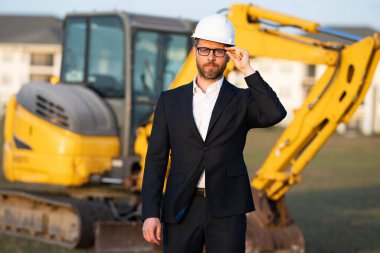 İnşaat alanında mimar. Modern ev inşaatında kask ve takım elbiseli mimar. Güvenlik yeleği ve takım elbisesi olan bir mimar. Evin arka planında duran kendine güvenen mimar.