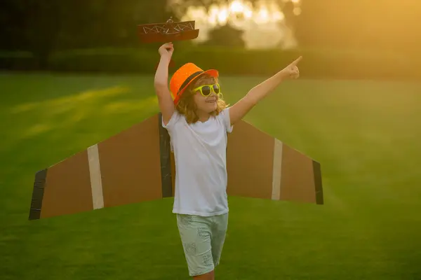 Kid pilot with paper wings or toy airplane having fun on meadow. Kid pilot aviator with with paper wings dreams of traveling in summer in park outdoor