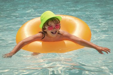 Yüzme havuzunda şişme yüzük takan bir çocuk. Çocuk turuncu şamandırayla yüzüyor. Su oyuncağı, çocuklar için sağlıklı açık hava sporları. Güneş gözlüklü moda yaz çocukları.