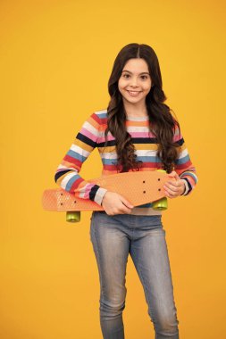 Teenagers lifestyle, casual youth culture. Teen girl with skateboard over isolated yellow studio background. Cool modern teenager in stylish clothes. Happy teenager, positive and smiling