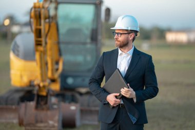 İnşaat işyeri sahibi. İnşaat alanında takım elbiseli ve şapkalı bir adam. İnşaat mühendisi kazıcının yanındaki evin önünde. Mimar, yönetici, yatırımcı, inşaat yöneticisi