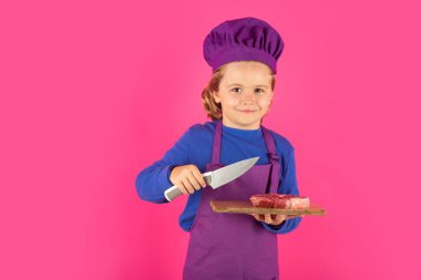 Çocuk aşçı et bifteği ve bıçakla kesme tahtasını tutuyor. Yemek pişiren çocuklar. Aşçı çocuk, aşçı kılığında.